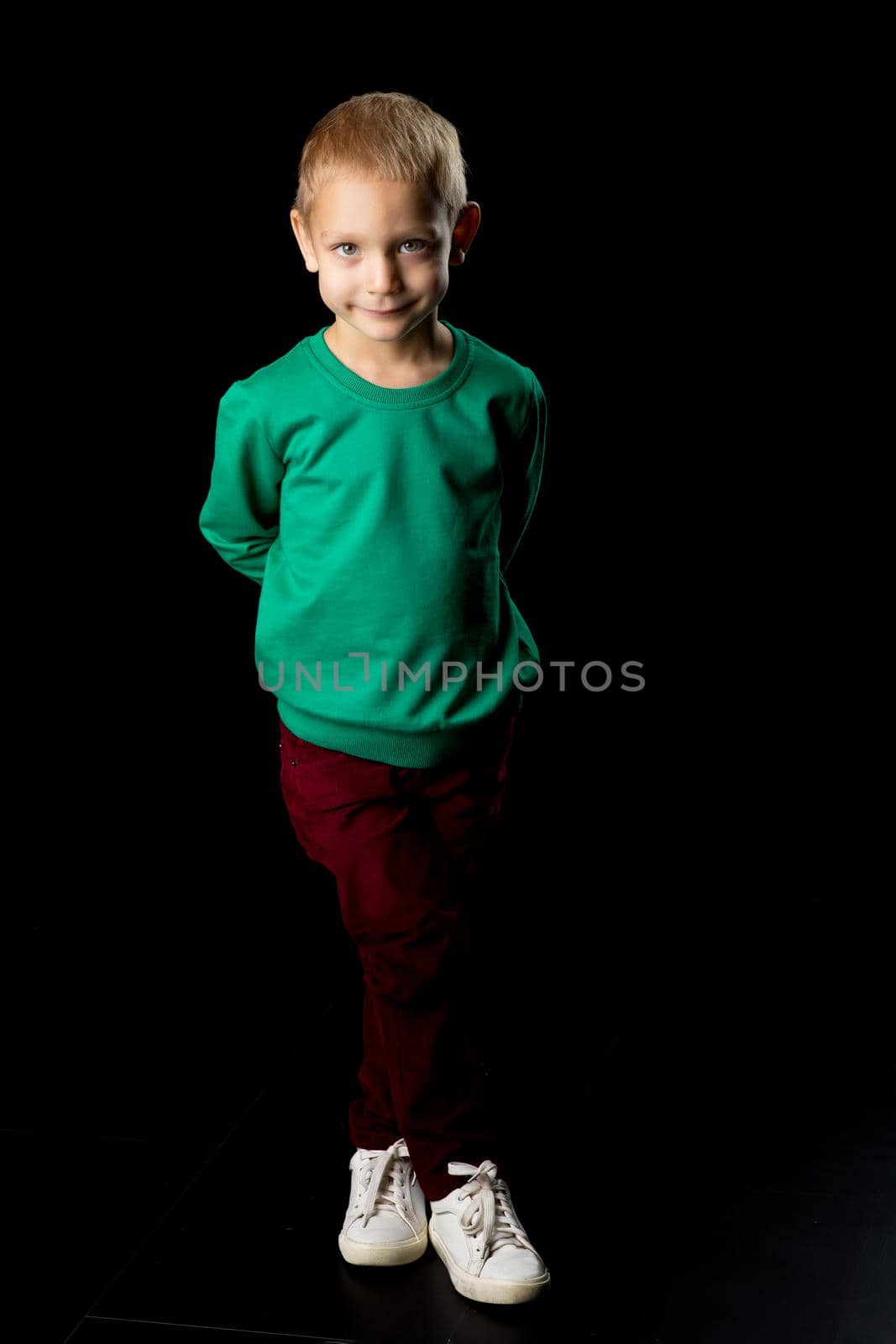 Cute boy posing in front of the camera in the studio, a charming boy in full growth in a green sweater. Portrait of a happy blond child, isolated on black background.