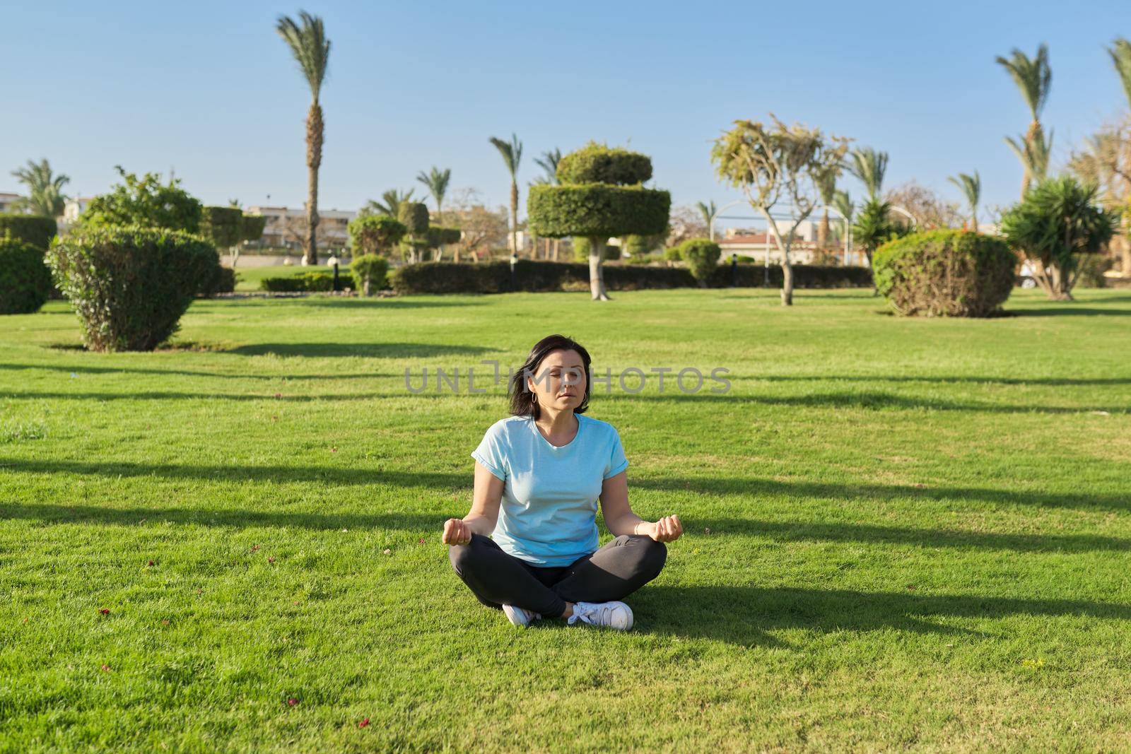 Middle aged woman sitting on grass in lotus position and meditating with closed eyes, sunny morning on a summer day. Meditation, relaxation, healthy lifestyle and body, mental health, people 40s