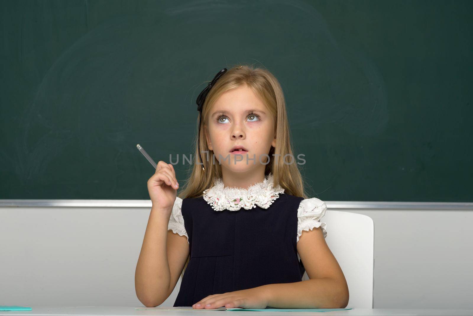 Girl thinking while sitting at desk at lesson. Beautiful blonde girl elementary school student posing on background of blackboard in classroom. School and education concept