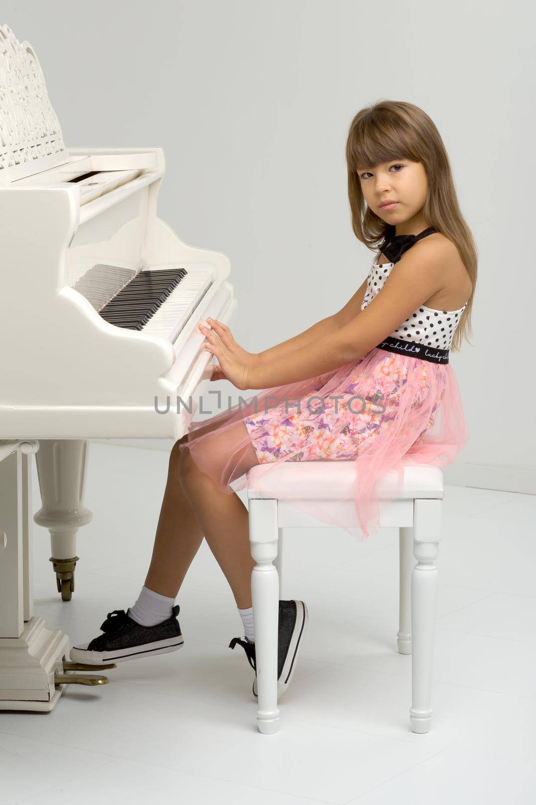 Young girl posing at white grand piano. Adorable girl sitting at at musical instrument. School of arts, music lesson concept. Portrait of preteen child in nice dress looking at camera