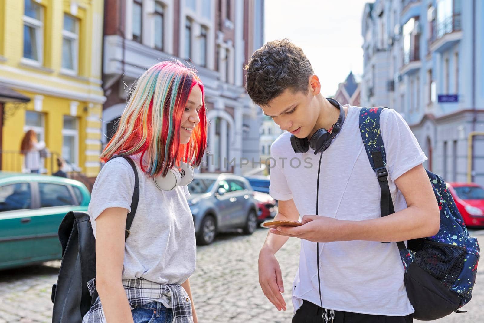Teenagers students guy and girl with backpacks and headphones look in smartphone screen, talk, city street background. High school and college, communication, youth concept