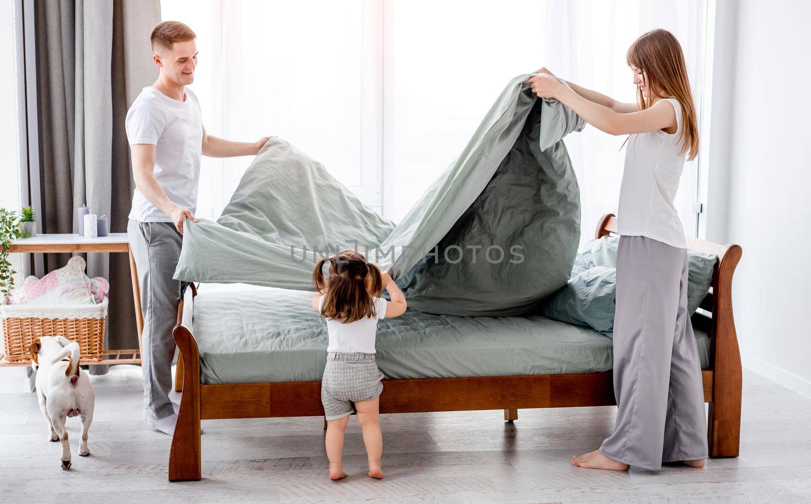 Young parents make a bed and their little daughter looking at them. Beautiful family morning with dog. Mother, father with their child and pet in sunny room