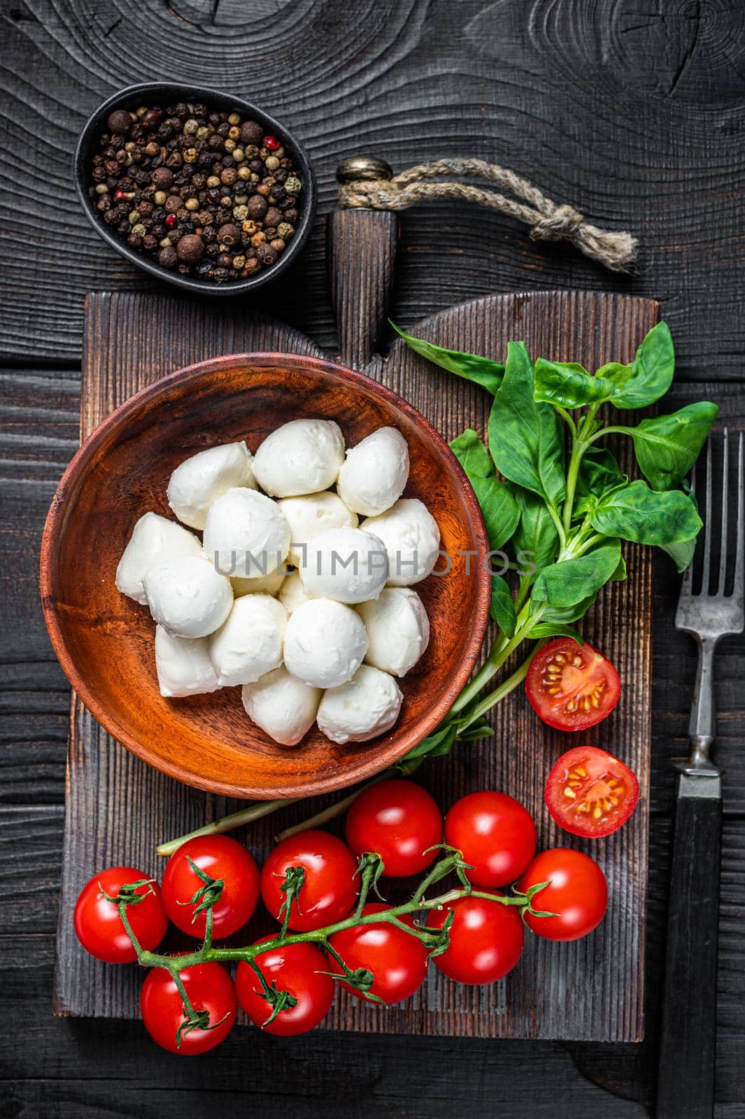 Italian mini Mozzarella cheese balls, basil and tomato cherry ready for cooking Caprese salad. Black wooden background. Top view by Composter