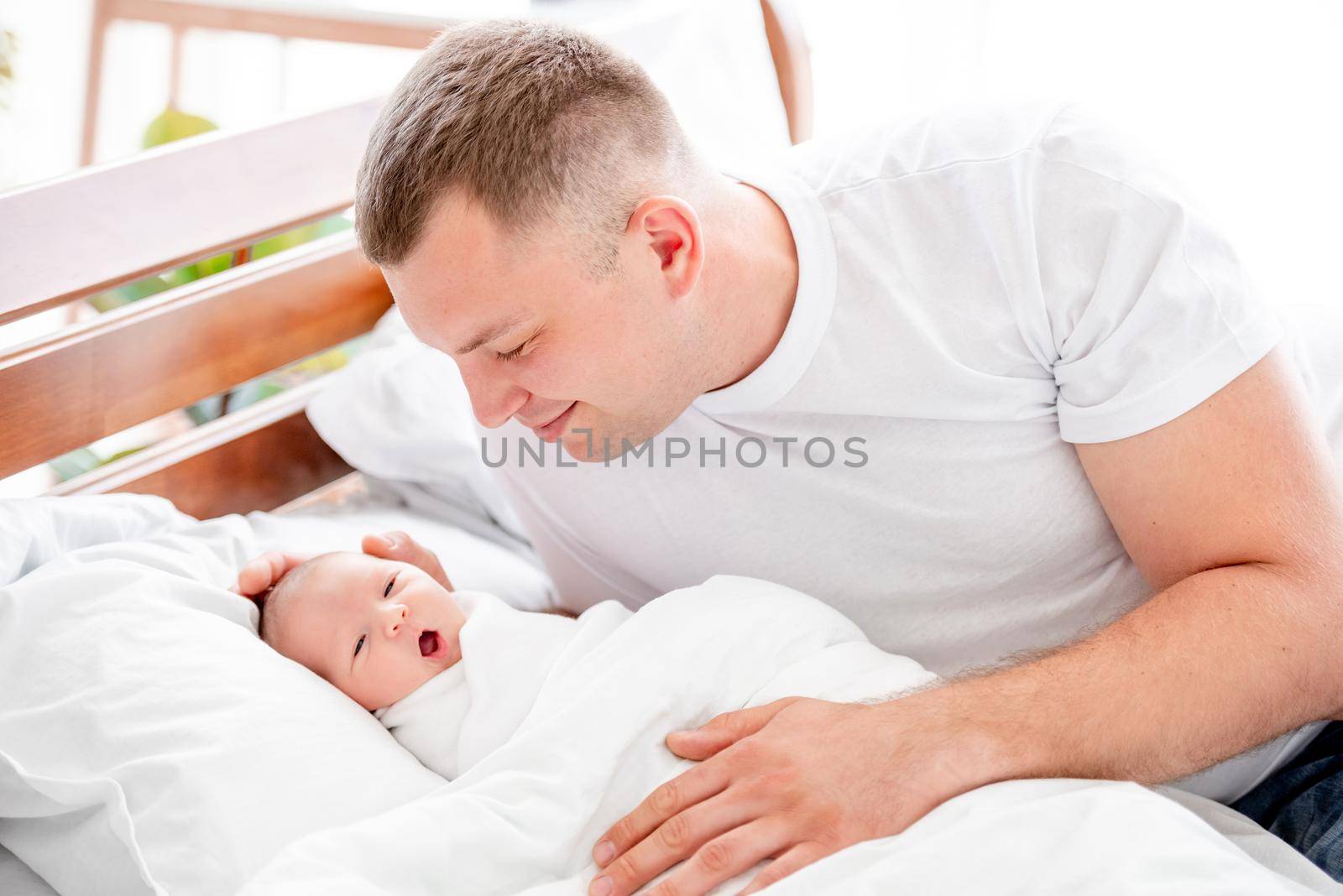 Young father sitting in the bed and looking with tenderness at his newborn baby daughter. Smiling parent dad and infant child girl at home with daylight