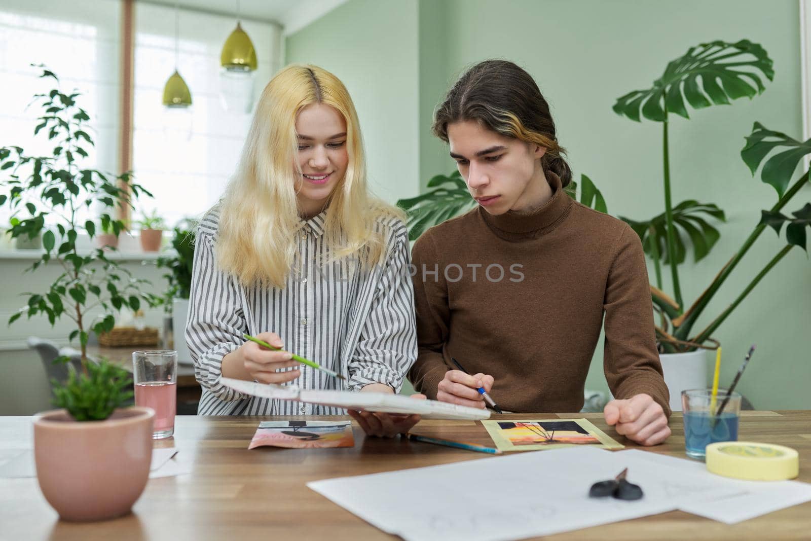 Teenagers painting with watercolors. Couple of guy and girl sitting at table with paints, brushes, drawings. Happy teens talking laughing creative. Creativity, hobby, leisure concept