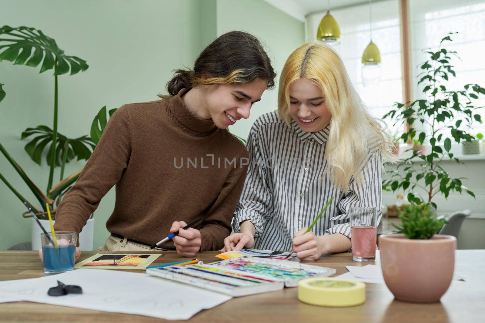 Teenagers painting with watercolors. Couple of guy and girl sitting at table with paints, brushes, drawings. Happy teens talking laughing creative. Creativity, hobby, leisure concept