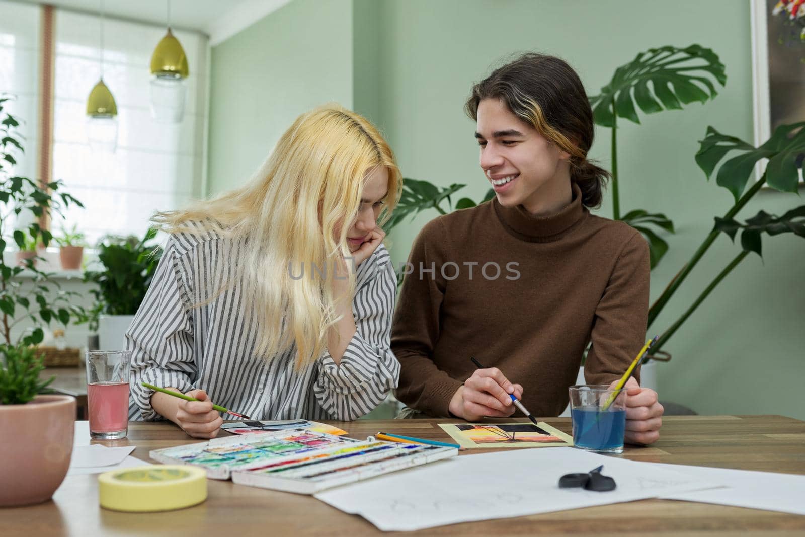 Teenagers painting with watercolors. Couple of guy and girl sitting at table with paints, brushes, drawings. Happy teens talking laughing creative. Creativity, hobby, leisure concept