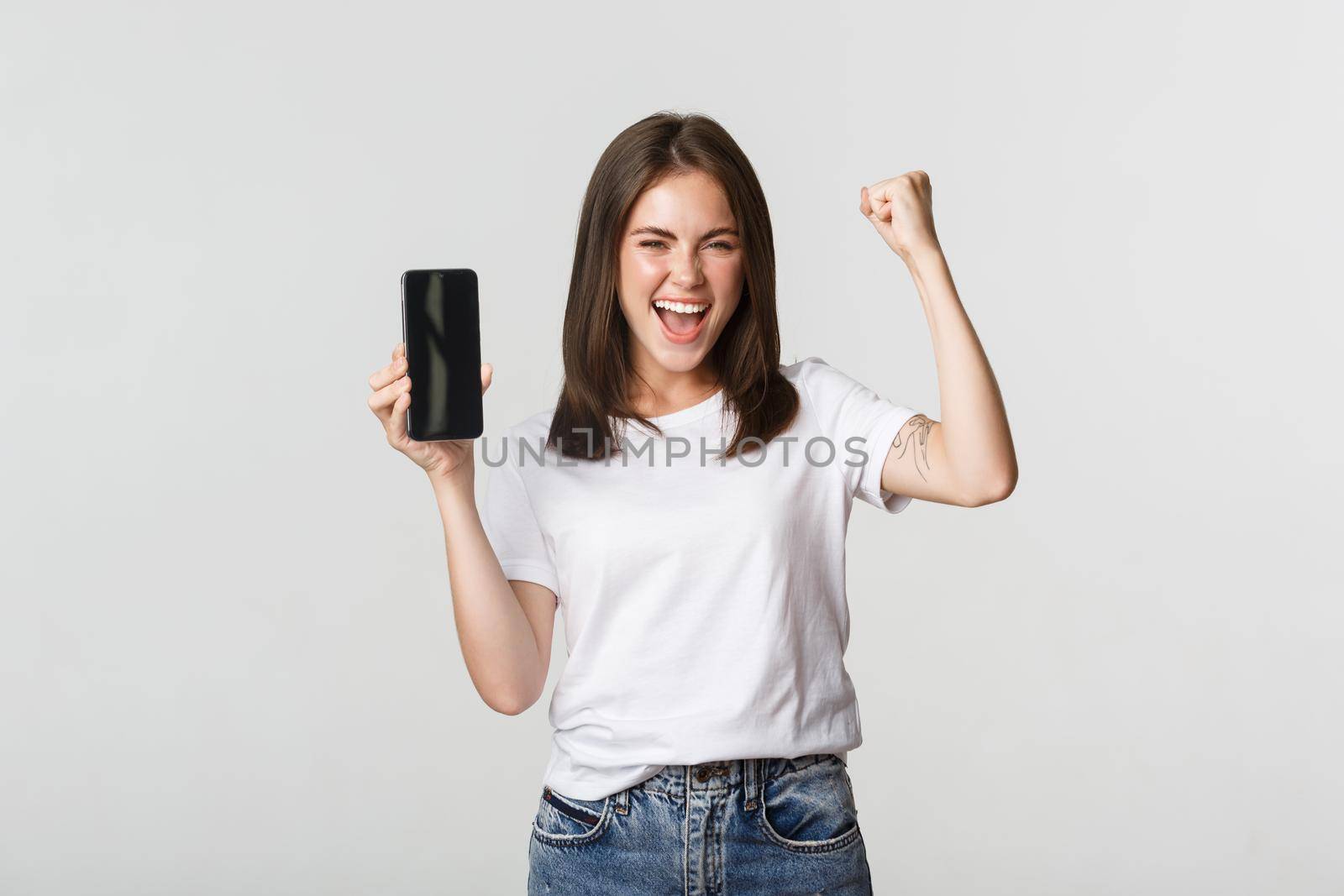 Rejoicing beautiful girl showing smartphone screen and dancing like champion.