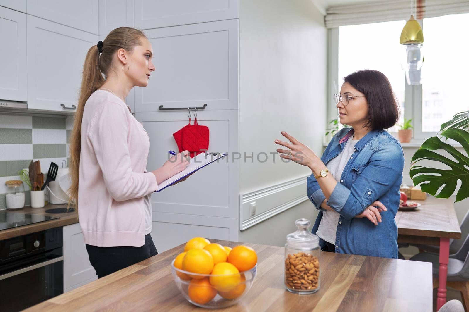 Poll, interview, monitoring, visitor young woman social worker talking and interrogating mature woman housewife, home kitchen interior background
