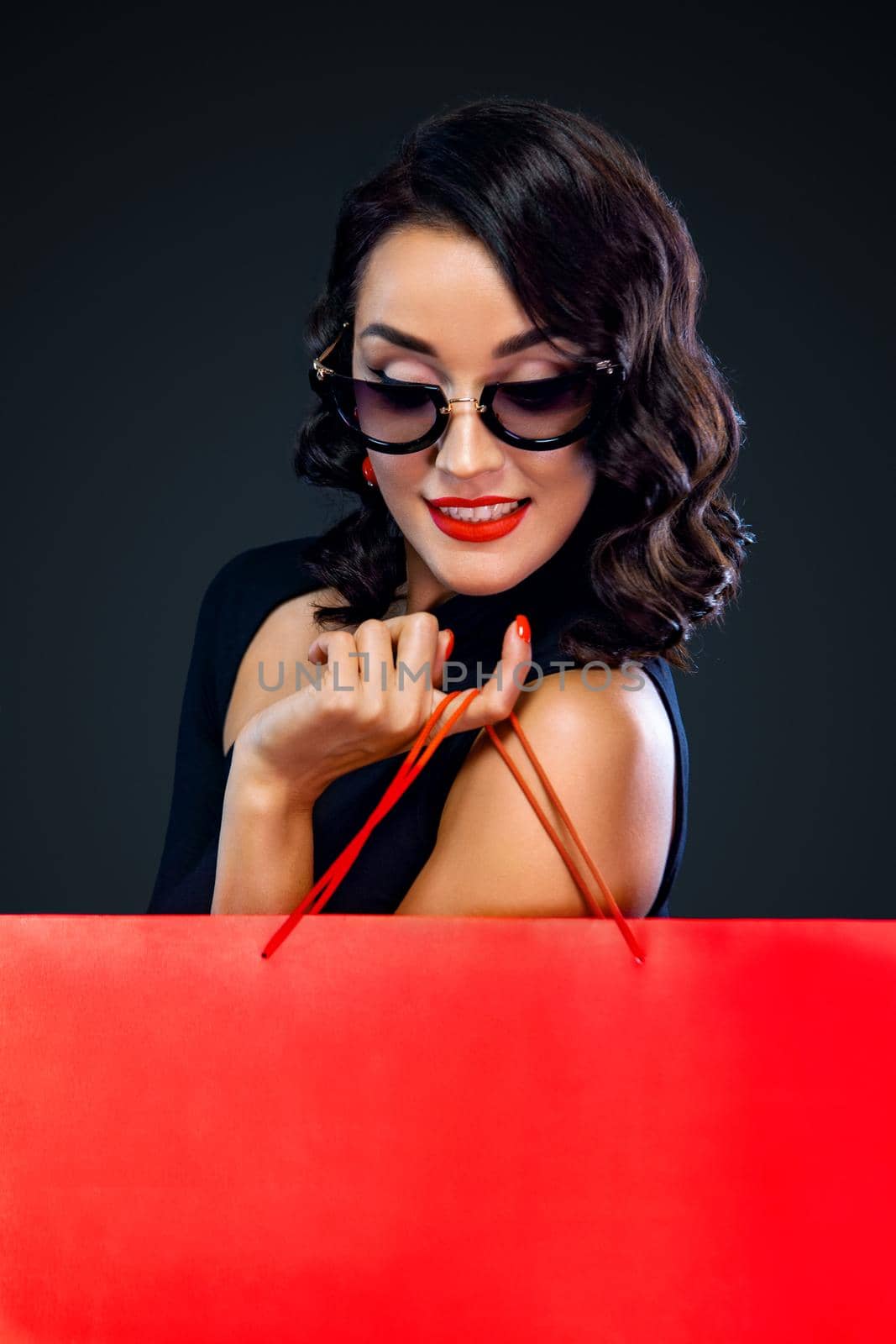 Black Friday sale concept for shop. Shopping woman in sunglasses holding red bag isolated on dark background. by MikeOrlov