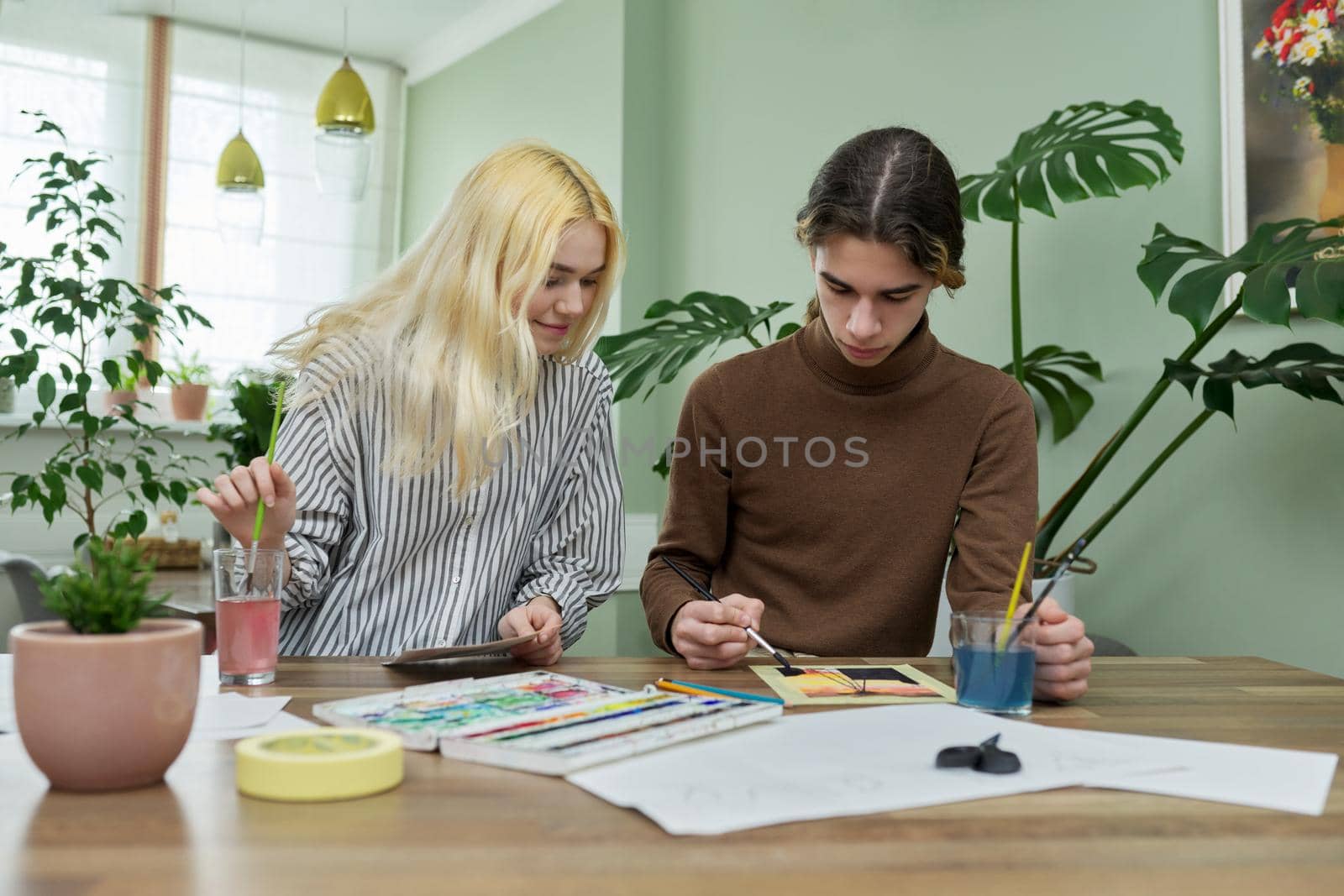 Teenagers painting with watercolors. Couple of guy and girl sitting at table with paints, brushes, drawings. Happy teens talking laughing creative. Creativity, hobby, leisure concept