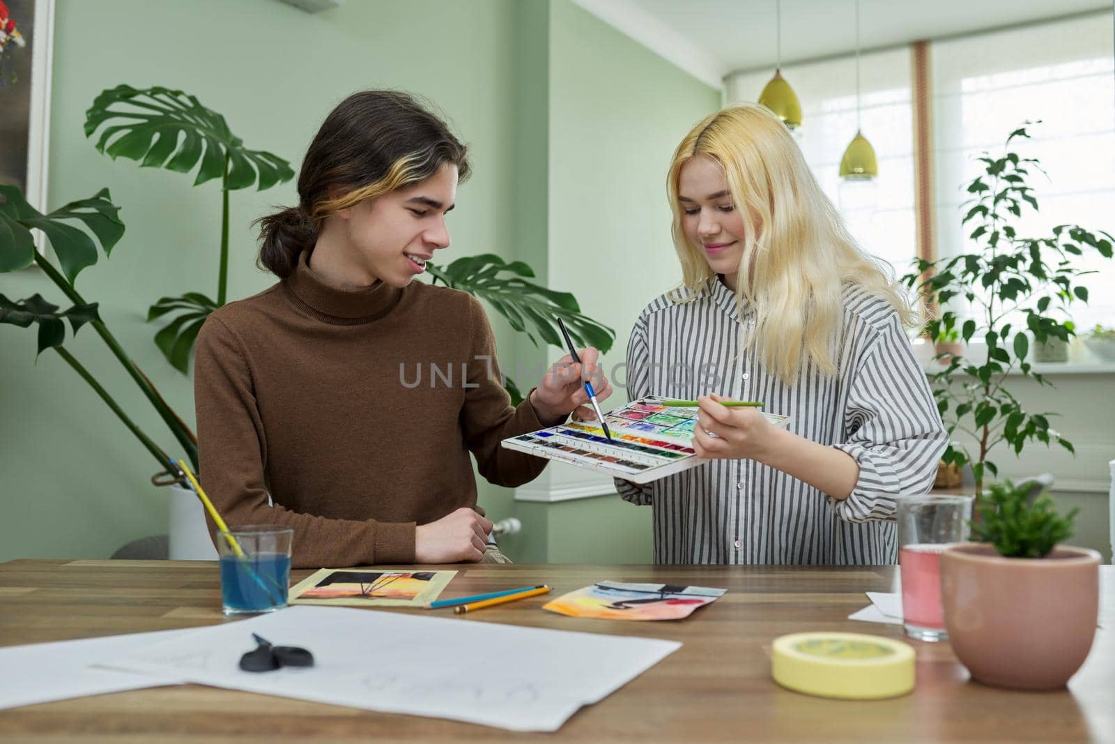 Teenagers painting with watercolors. Couple of guy and girl sitting at table with paints, brushes, drawings. Happy teens talking laughing creative. Creativity, hobby, leisure concept