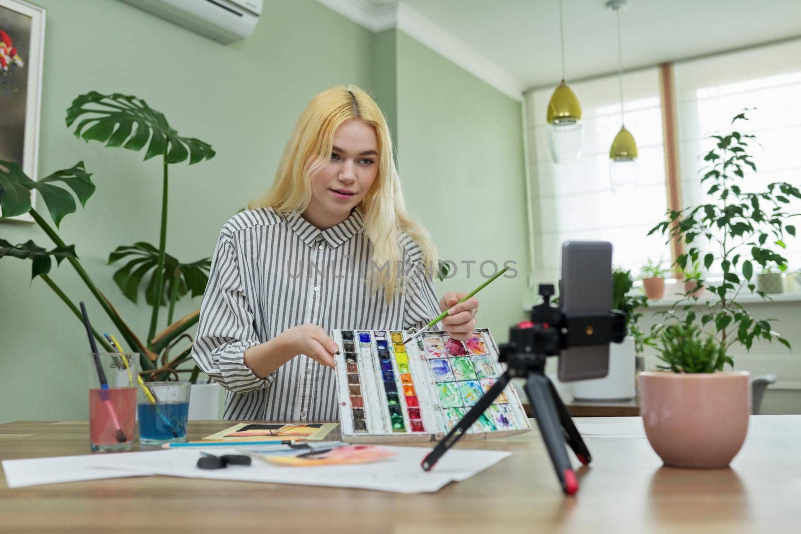 Teenage girl recording video drawing lesson using smartphone on tripod, showing watercolor paints. Hobby, leisure, technology, online learning, creativity, teenagers concept