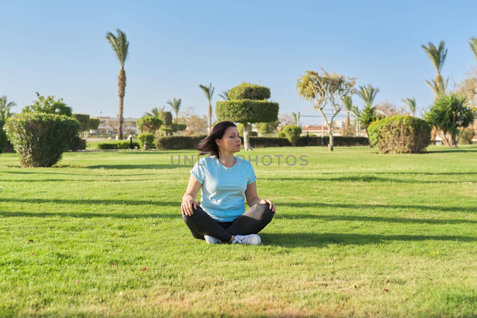 Middle aged woman sitting on grass in lotus position and meditating with closed eyes by VH-studio