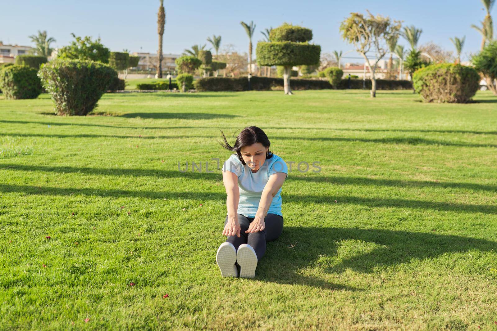 Middle-aged woman doing outdoor sports, 40s female sitting on grass on sunny summer morning. Healthy lifestyle, healthy body, mature people concept