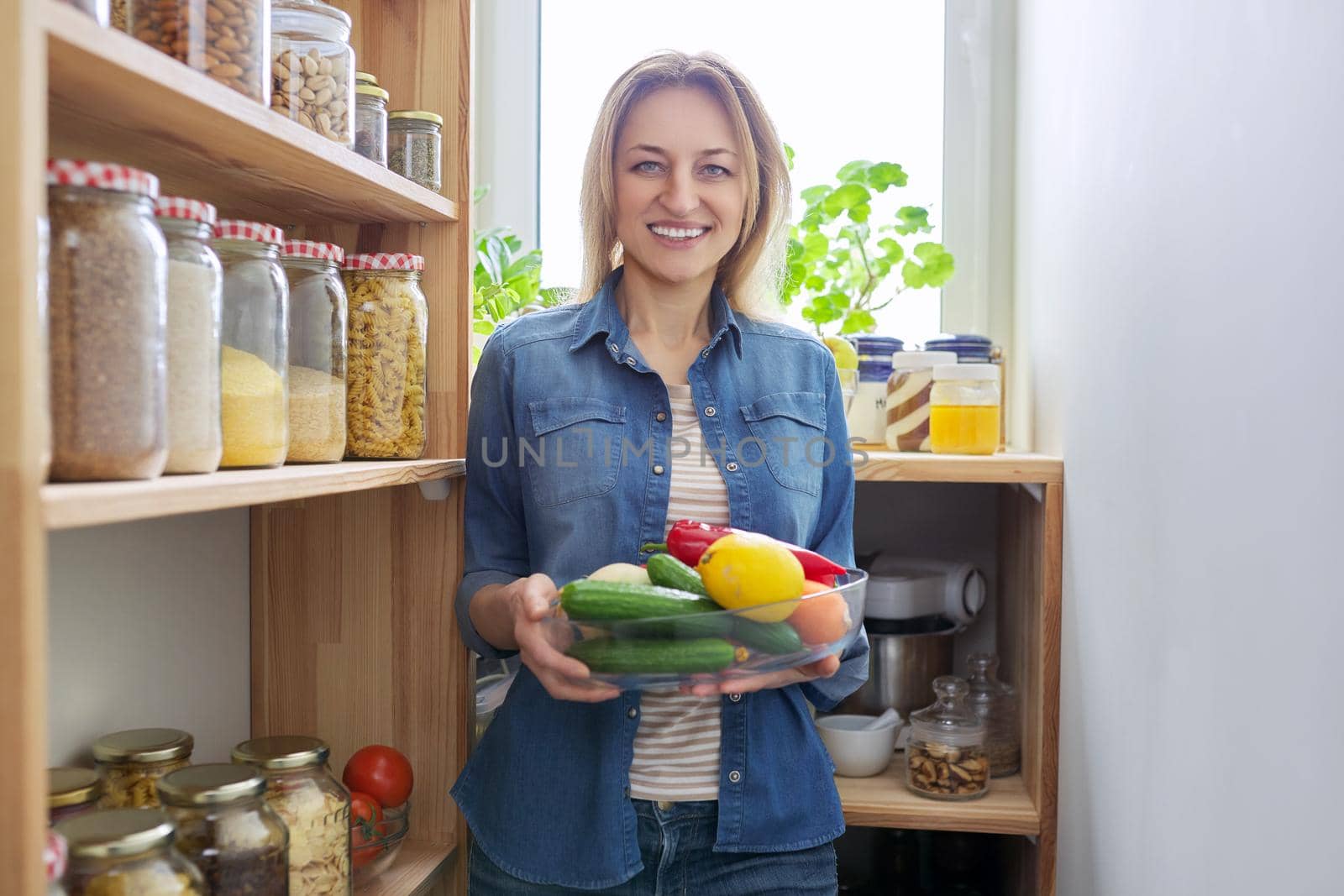 Portrait of a middle-aged woman in the kitchen in the pantry with a bowl of vegetables in her hands. by VH-studio