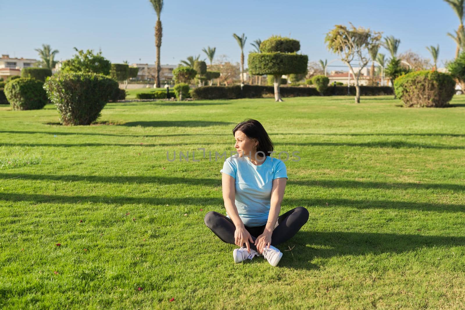 Middle-aged woman doing outdoor sports, 40s female sitting on grass on sunny summer morning by VH-studio