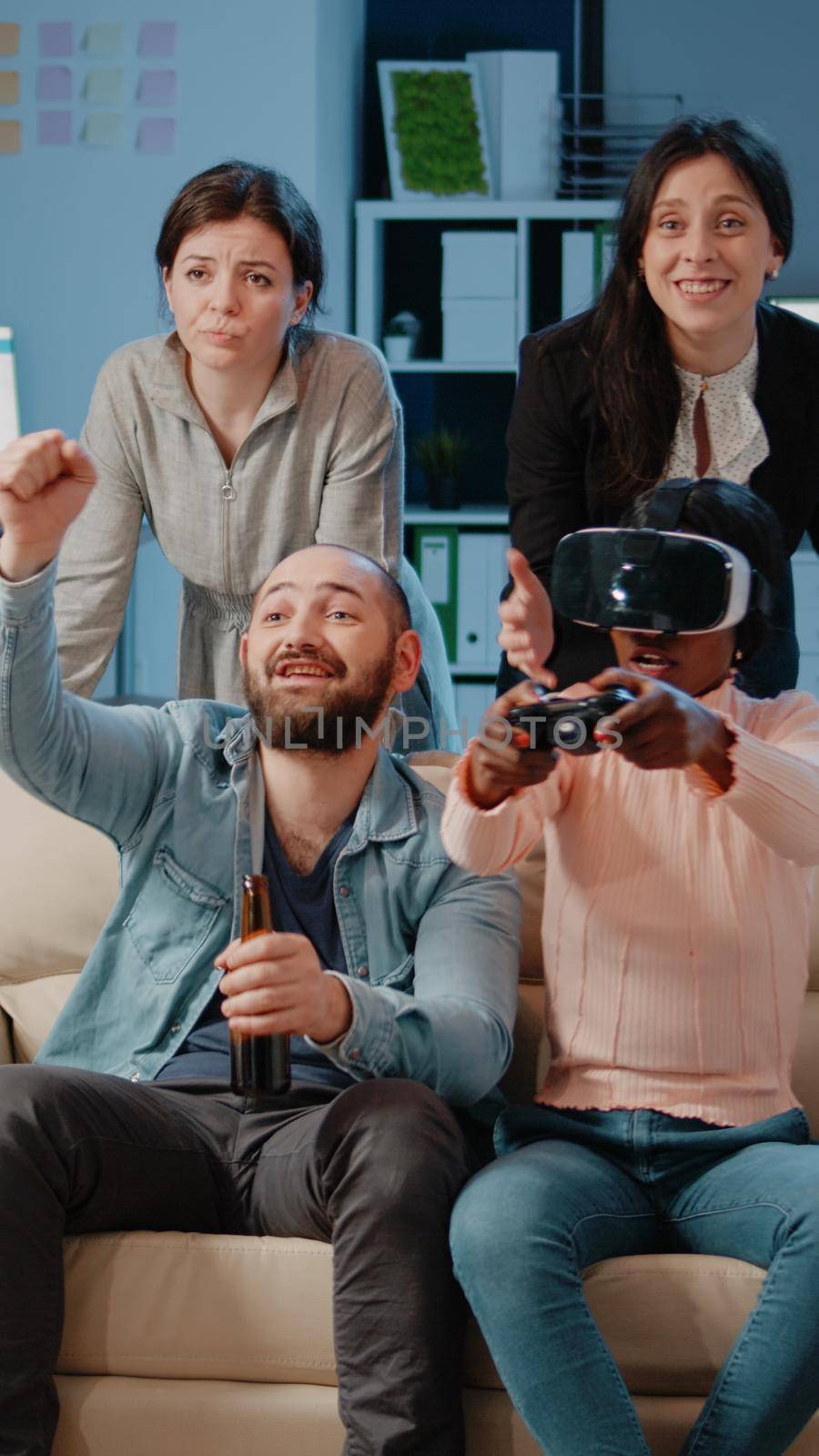 African american woman using vr glasses and controller to play video games while colleagues cheering for entertainment after work. Coworkers playing with technology to do fun activity