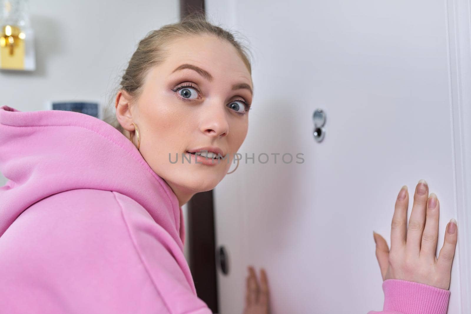 Young woman looking through the peephole of front door in apartment. Emotion of surprise on female's face