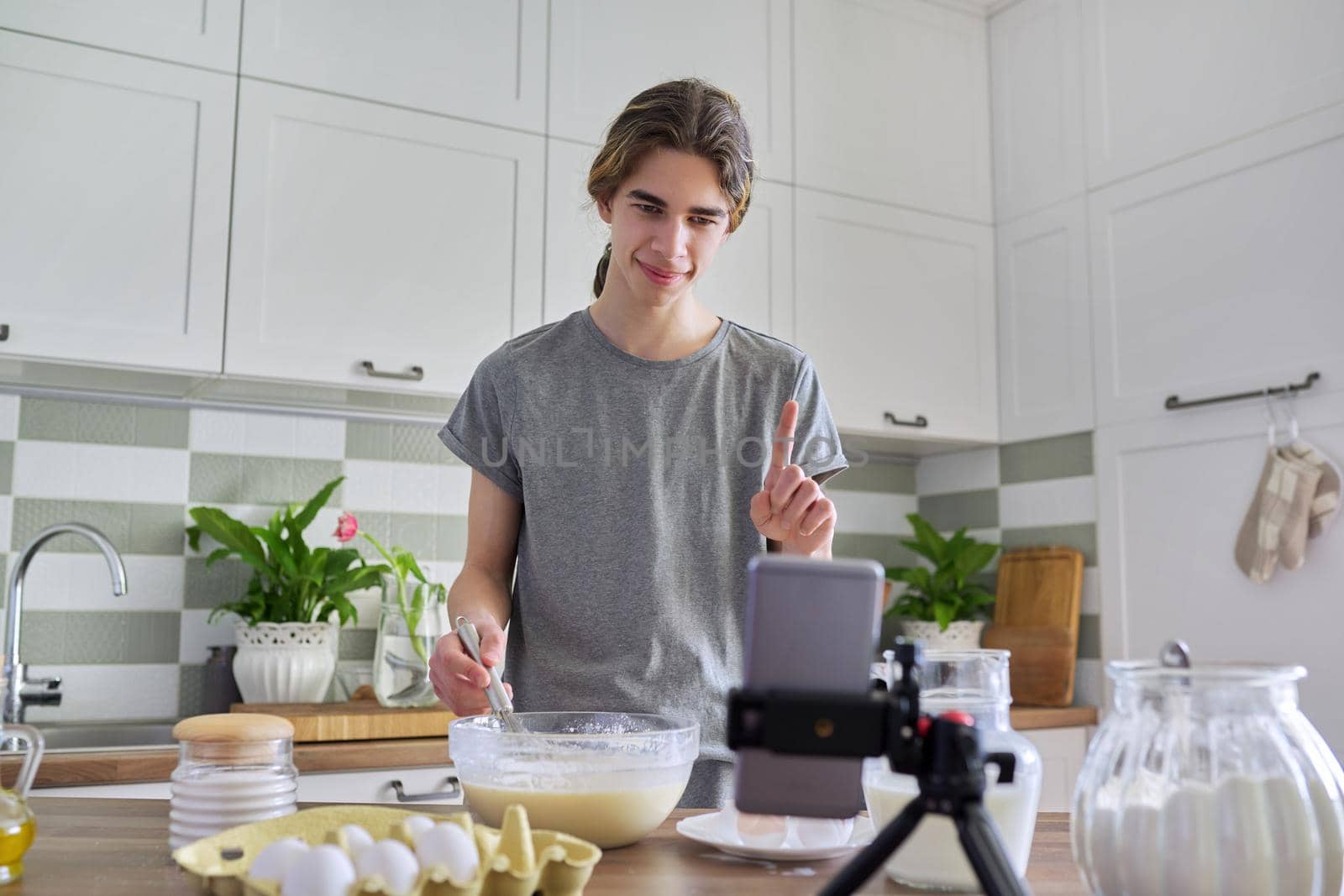 Male teenager cooking pancakes talking on video communication on smartphone by VH-studio