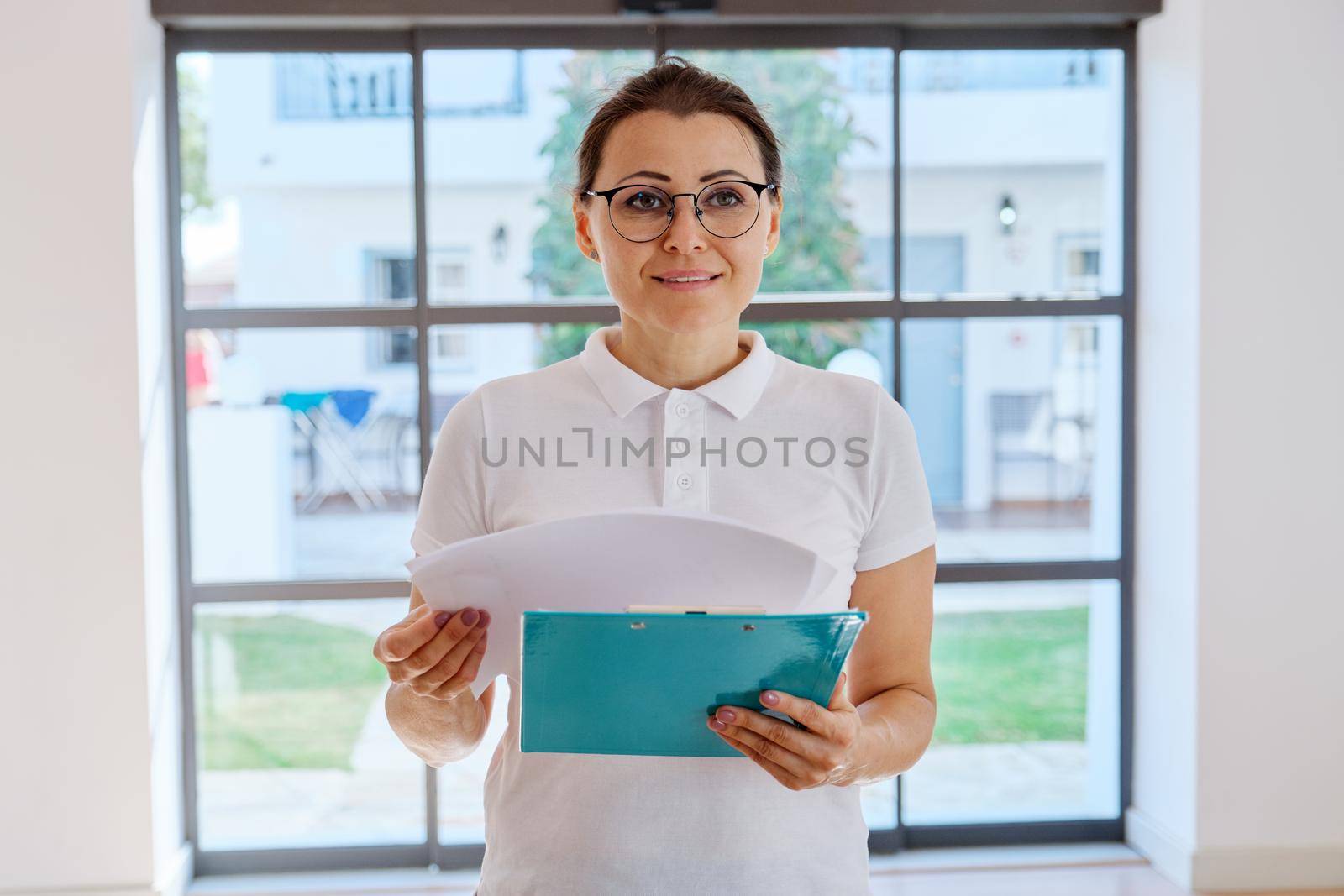 Business woman portrait with clipboard in hand. Middle-aged female wearing white T-shirt glasses, inside office hall. People, workers, profession, staff, occupation, administration concept