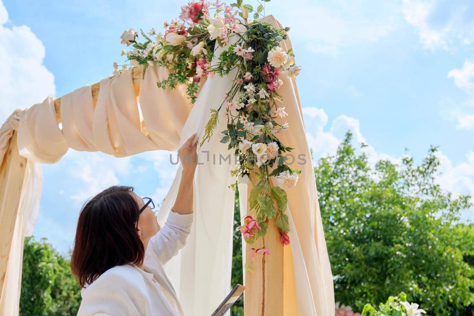 Organization, preparation for holidays, wedding, event. Decorating arch with textiles with flowers and plants. Woman organizer, owner, with digital tablet near wedding arch