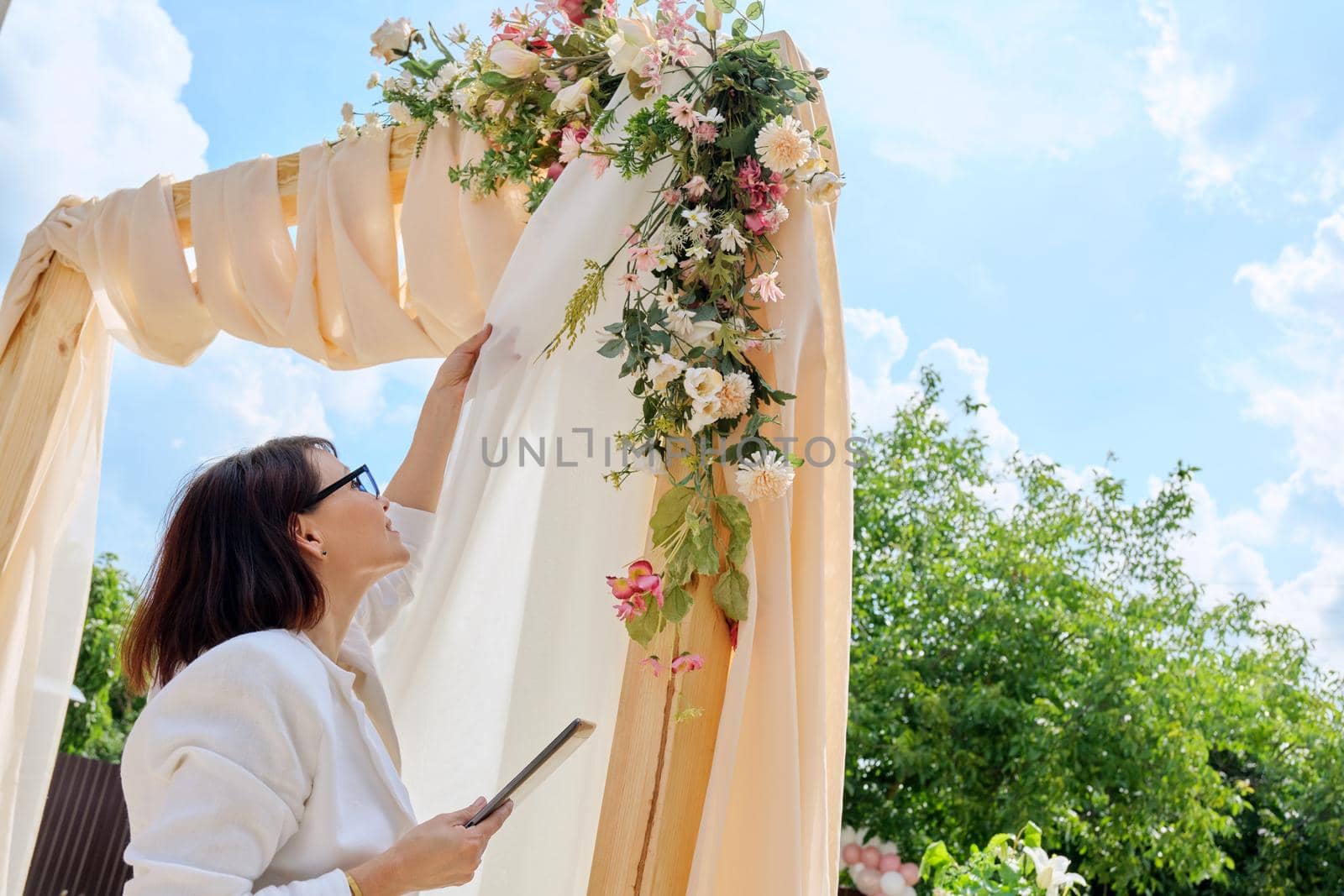 Organization, preparation for holidays, wedding, event. Decorating arch with textiles with flowers and plants. Woman organizer, owner, with digital tablet near wedding arch