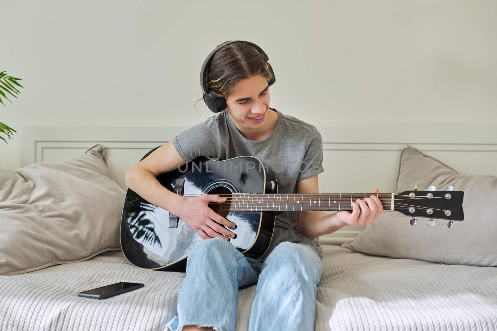 Male teenager in headphones sitting at home on bed with acoustic guitar and smartphone. Hipster guy using smartphone to learn music, study songs, record his music. Creativity, hobby, teens concept