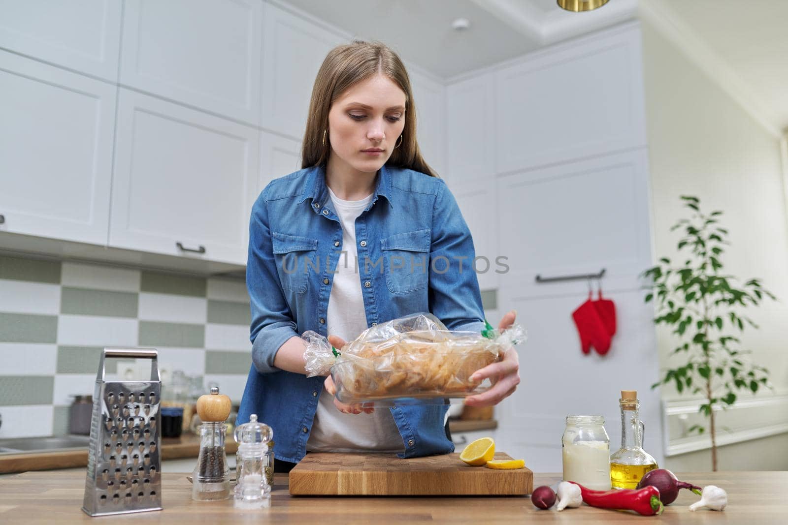 Young woman cooking baked chicken in baking sleeve with spices, kitchen interior background, cooking at home concept