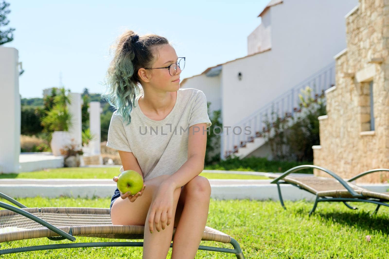 Teenage girl sitting outdoors in garden lounger on green lawn holding an apple in her hand, sunny summer day. Teens, holidays, rest, fruits, healthy food