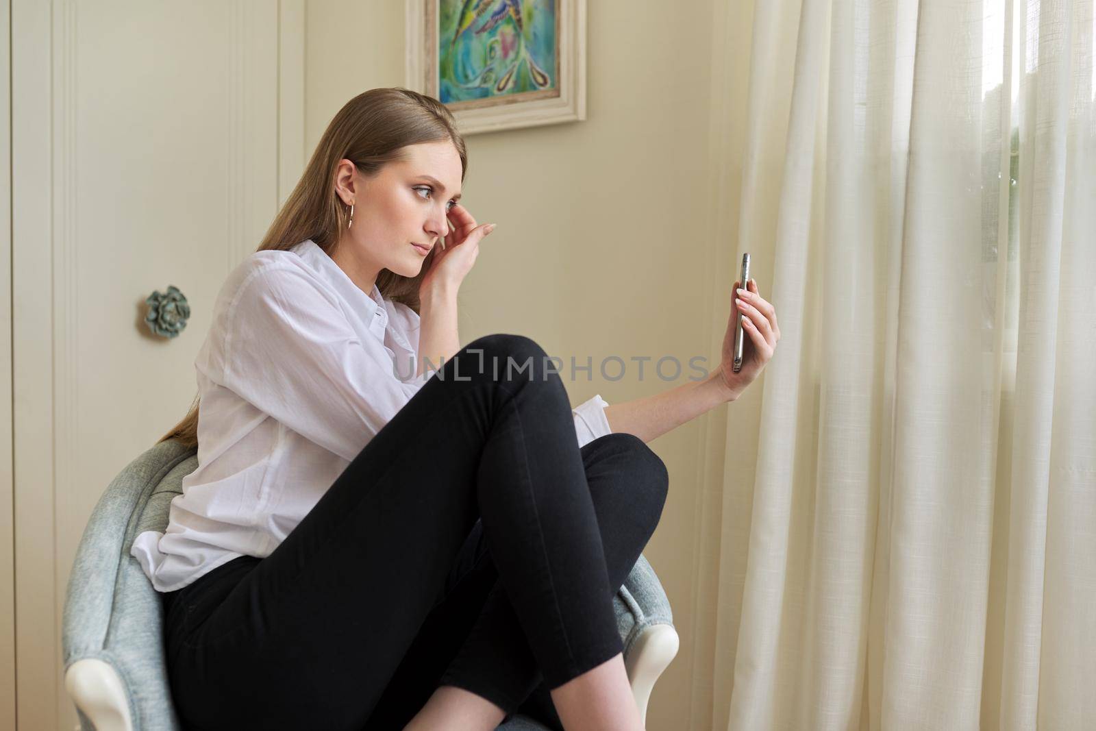 Young beautiful blonde sitting at home in chair, serious talking using video communication on smartphone. Leisure, woman relaxing looking at phone screen