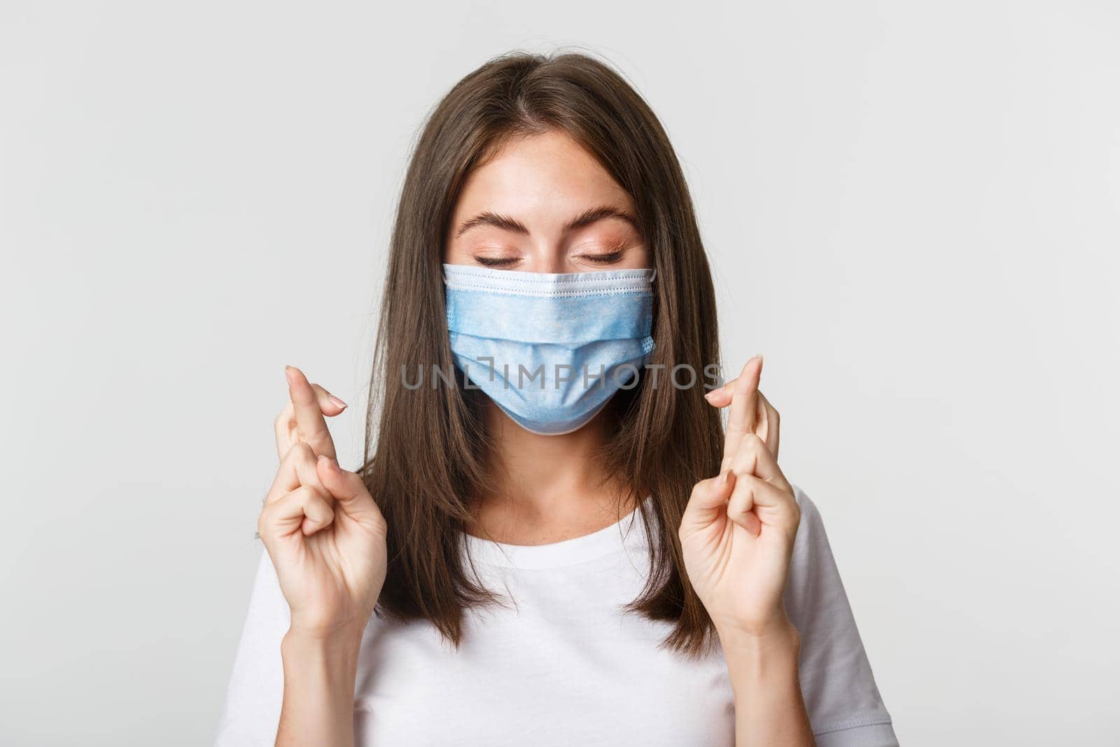 Covid-19, health and social distancing concept. Close-up of hopeful pretty girl in face mask close eyes and cross fingers good luck, making wish by Benzoix