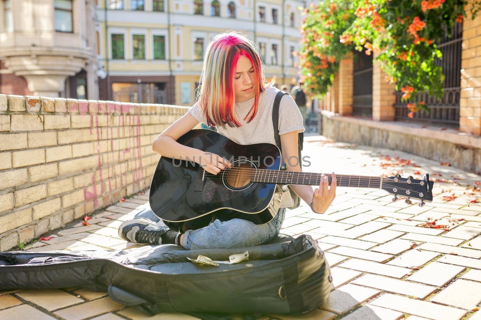 Creative trending female teenager musician playing acoustic guitar sitting on city street sidewalk with guitar case and cash