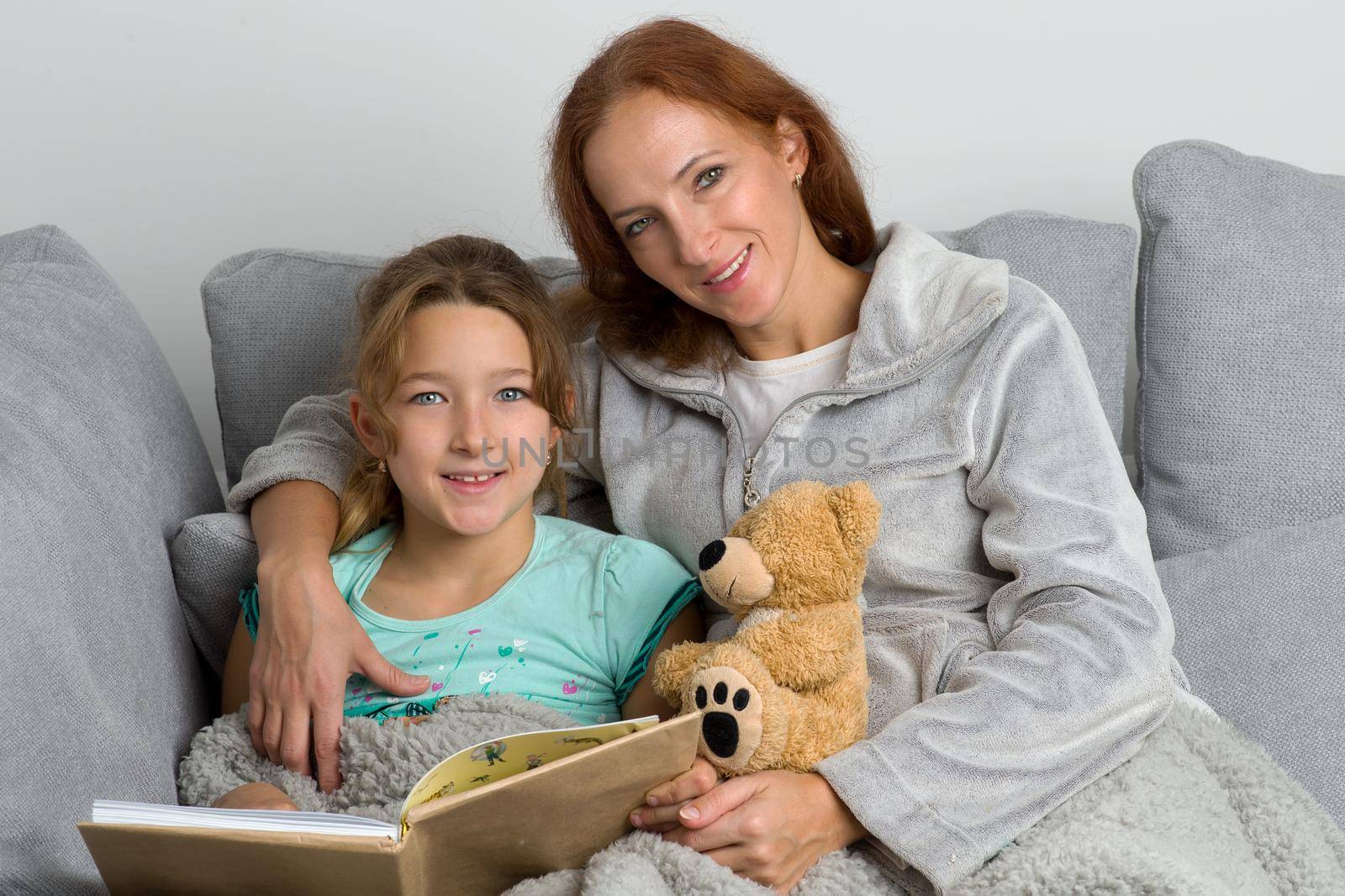 Mom sitting on couch with her daughter and reading book. Smiling loving mother hugging her daughter who sitting with teddy bear toy. Woman and child wearing indoor clothes sitting together