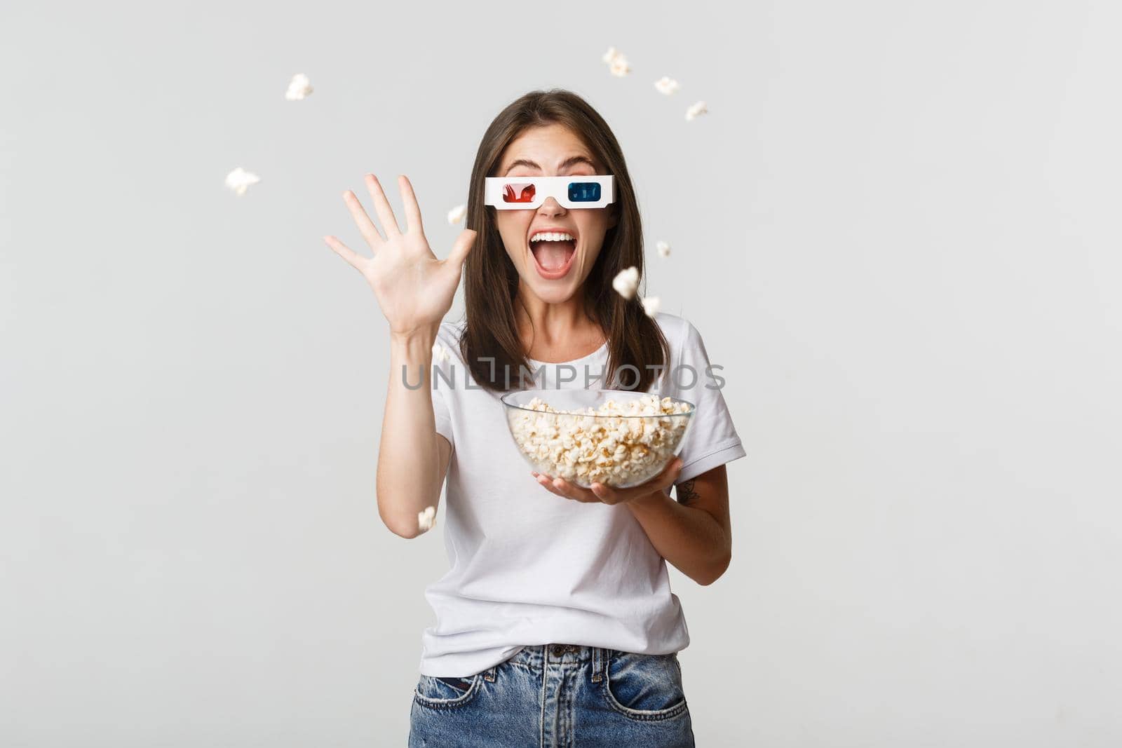 Joyful attractive young girl in 3d glasses, throwing popcorn at screen while watching movie.