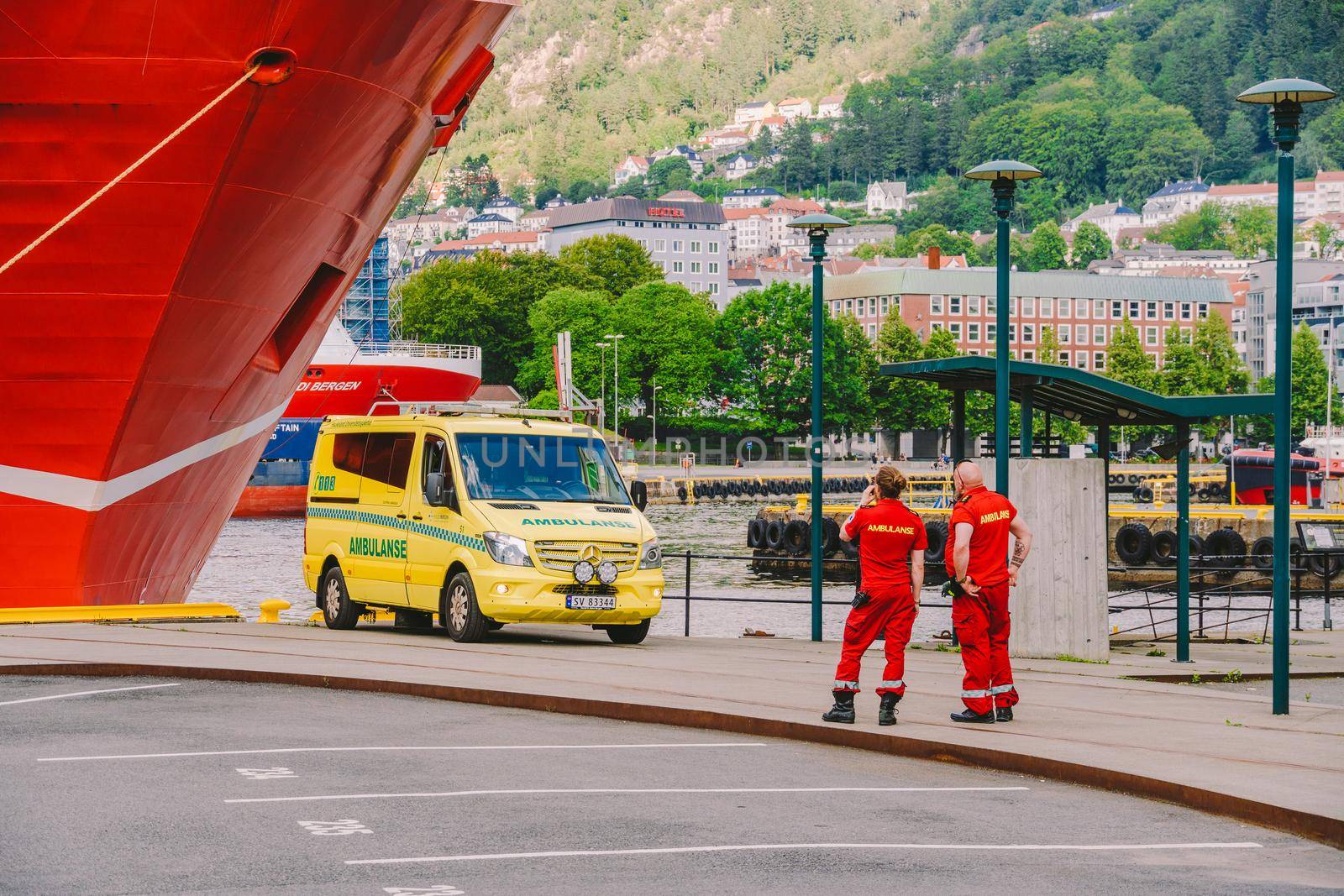 Two Norwegian paramedics in red uniforms are resting near an ambulance parked in a port near a large ship. Theme healthcare and medicine in Norway, Bergen July 28, 2019 by Tomashevska