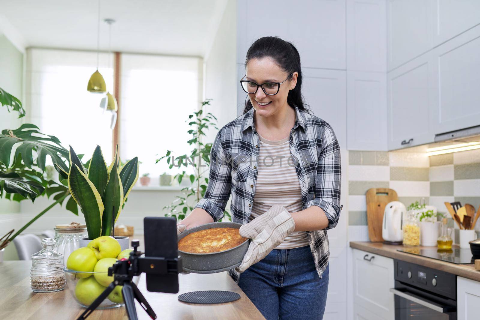 Food blog, woman blogger cooking apple pie at home, recording recipe on smartphone. Positive woman at home in kitchen talking online with friends family, using video call