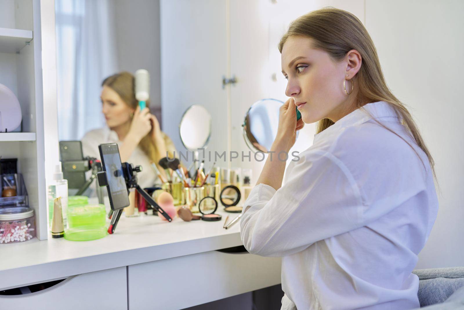 Beauty blog, young woman sitting at make-up table with mirror doing makeup hairstyle, styling her hair with brush, blonde female beauty blogger recording blog vlog on smartphone, online master class