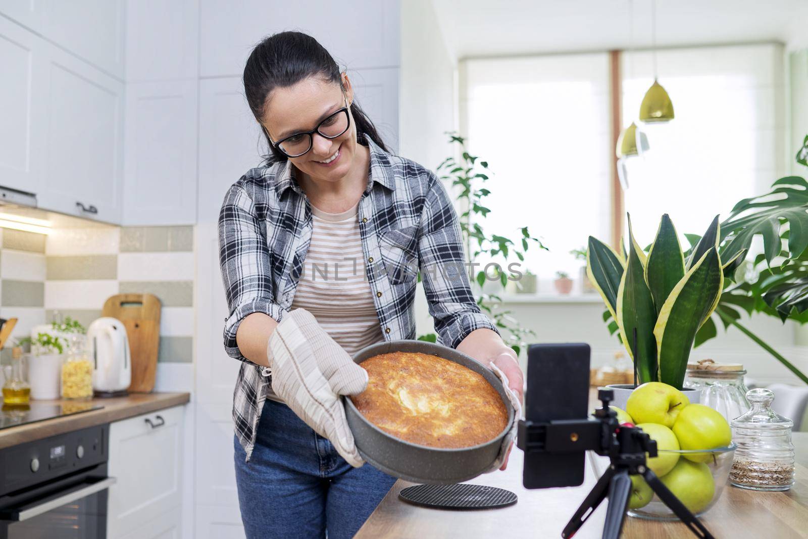 Food blog, woman blogger cooking apple pie at home, recording recipe on smartphone. Positive woman at home in kitchen talking online with friends family, using video call