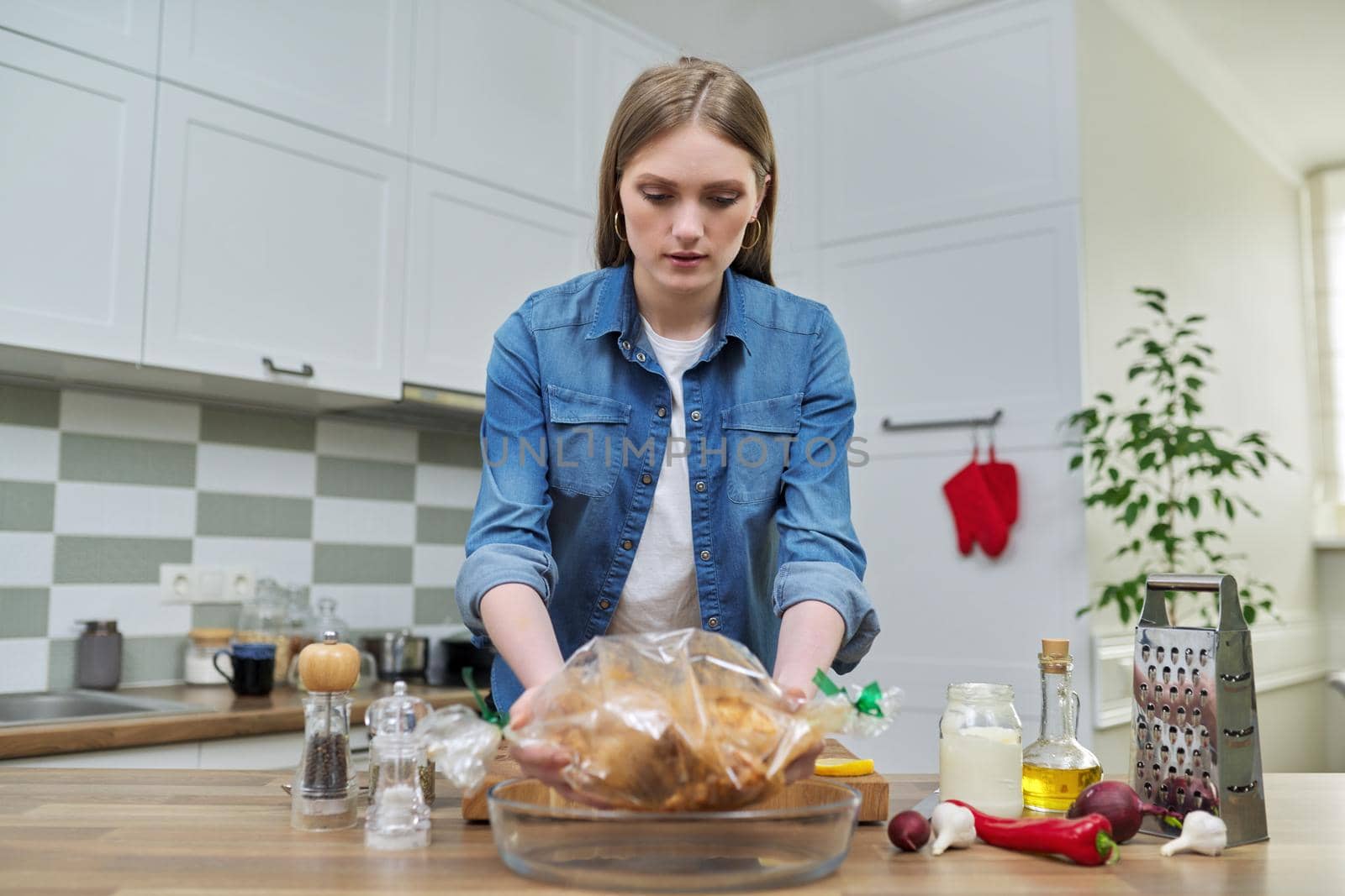 Young woman cooking baked chicken in baking sleeve with spices, kitchen interior background, cooking at home concept