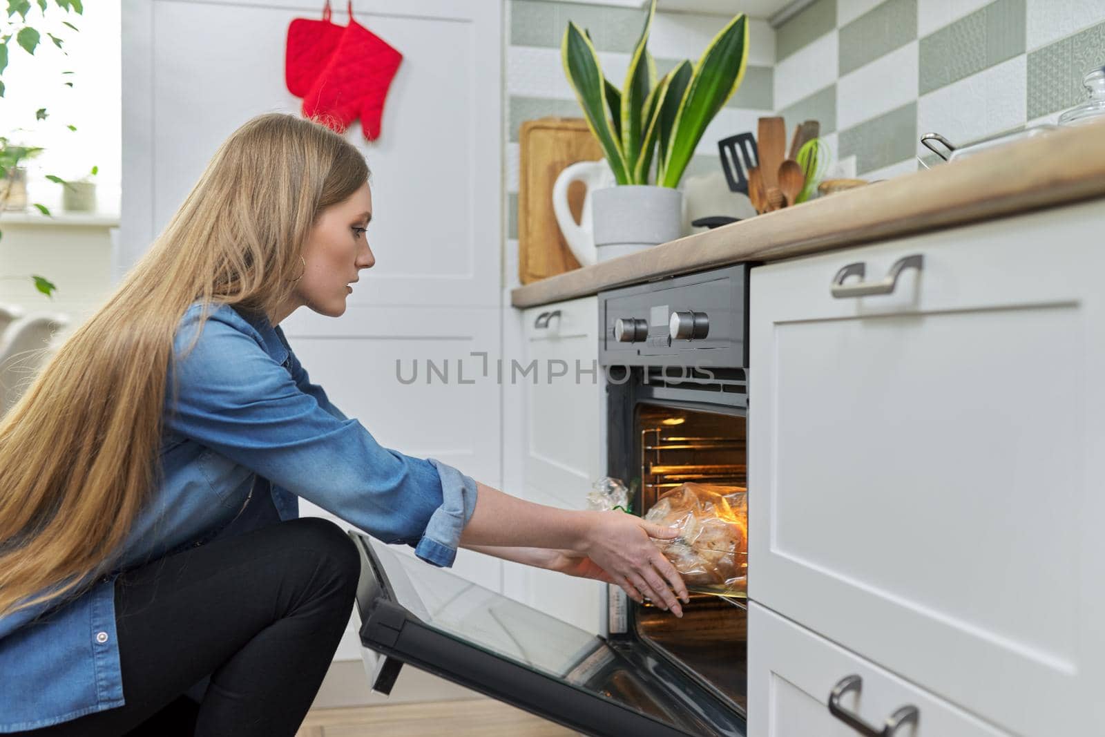 Cooking at home, roasting meat in oven, young woman putting marinated chicken in baking bag in oven, kitchen interior background