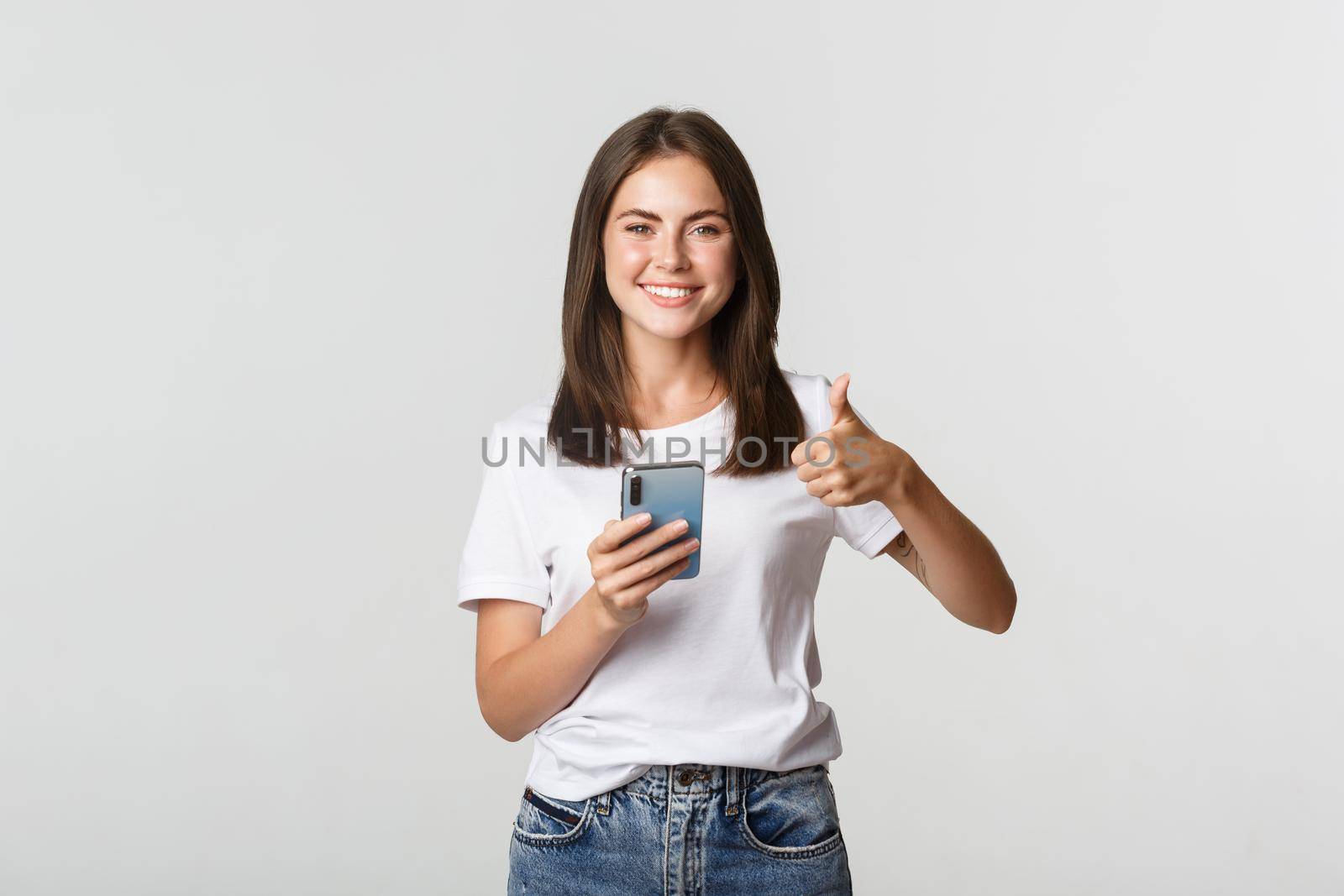 Satisfied smiling young woman showing thumbs-up while using smartphone.