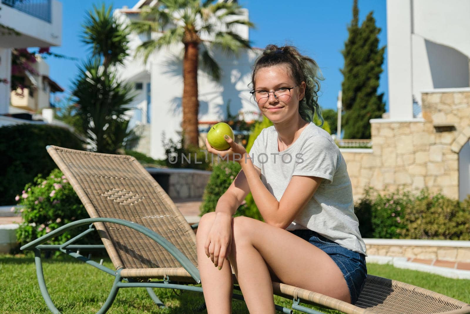 Girl teenager with green apple, resting sitting on sun lounger in garden, female holding tasty healthy apple in hand, sunny summer day grass background