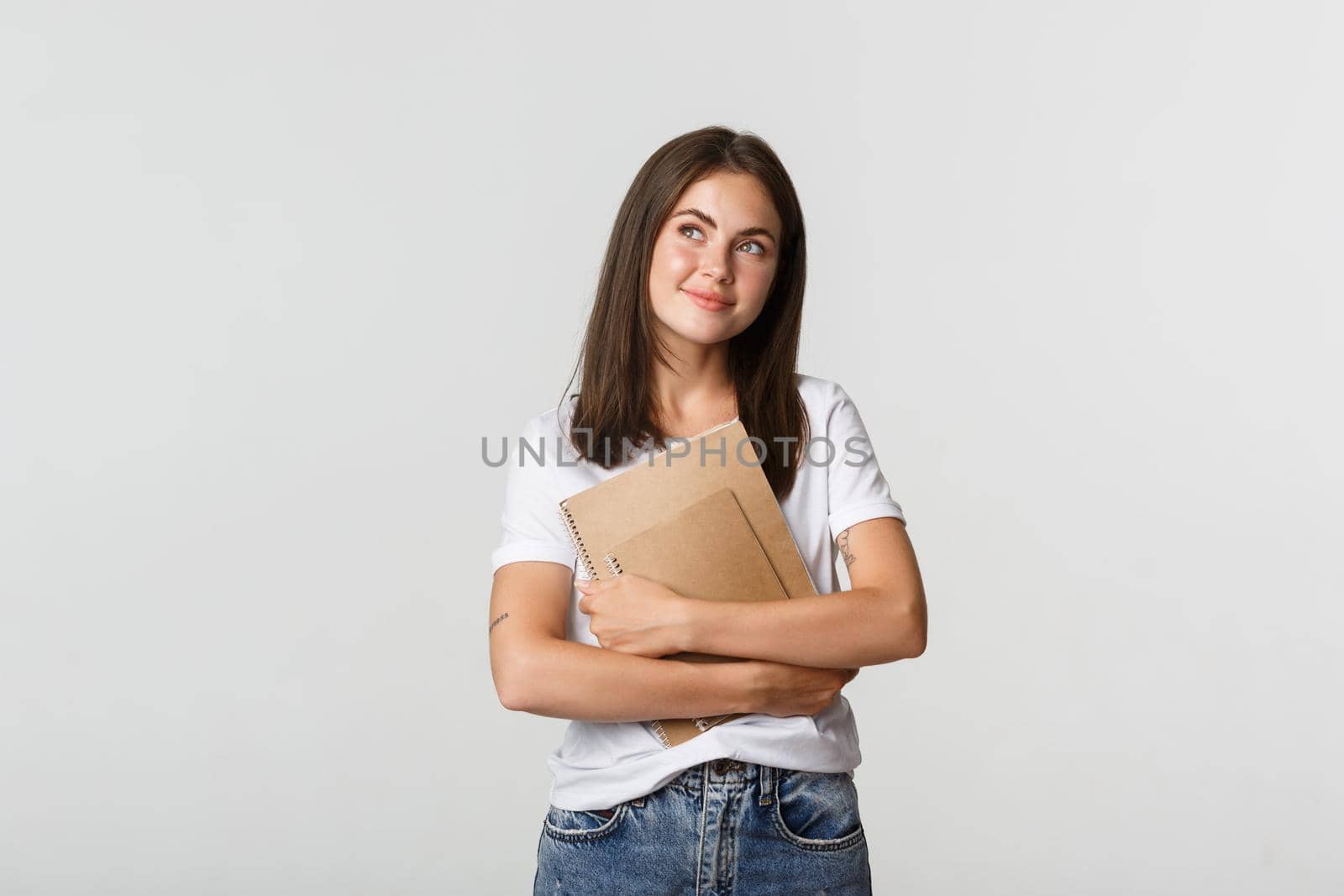 Portrait of smiling nostalgic female student looking upper left corner, holding notebooks.