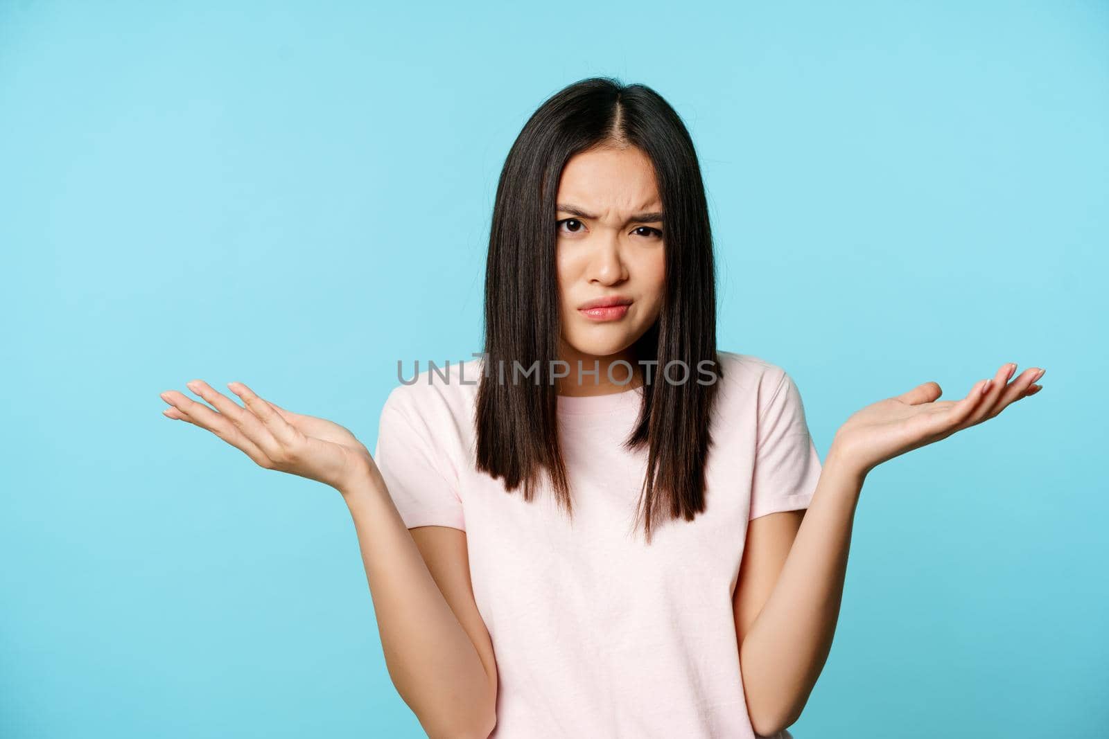 Confused and annoyed asian girl shrugging shoulders, furrow eyebrows and spread hands puzzled, cant understand smth, blue background.