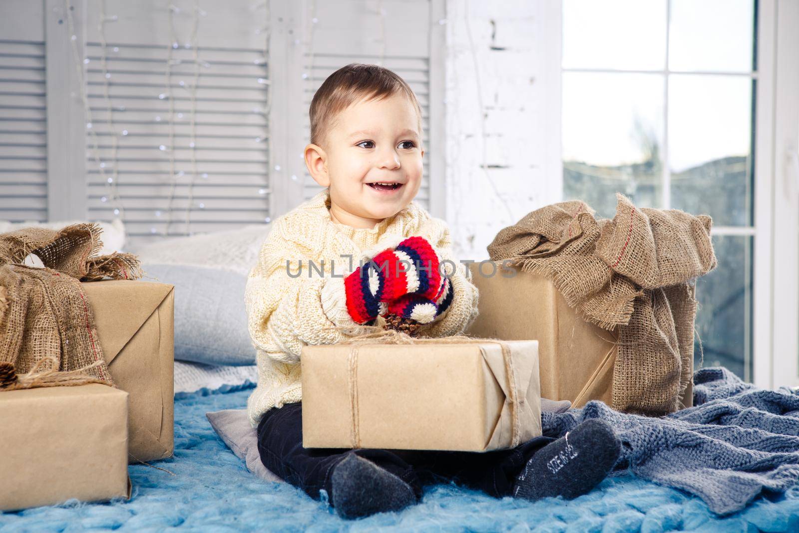 little funny playful boy a child sits on a bed on Christmas day with gift boxes in white wool knitted sweater and big bright mittens on it and laughs out loud. In interior there is a festive decor by Tomashevska
