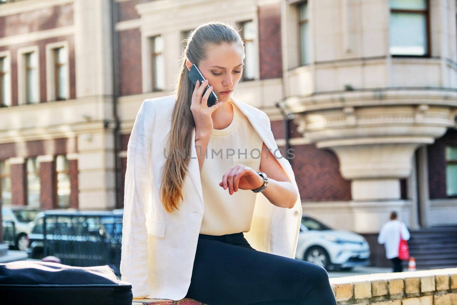 Young business woman in city looking at her wristwatch. Confident serious female talking on cell phone, sunset in the city, urban style background