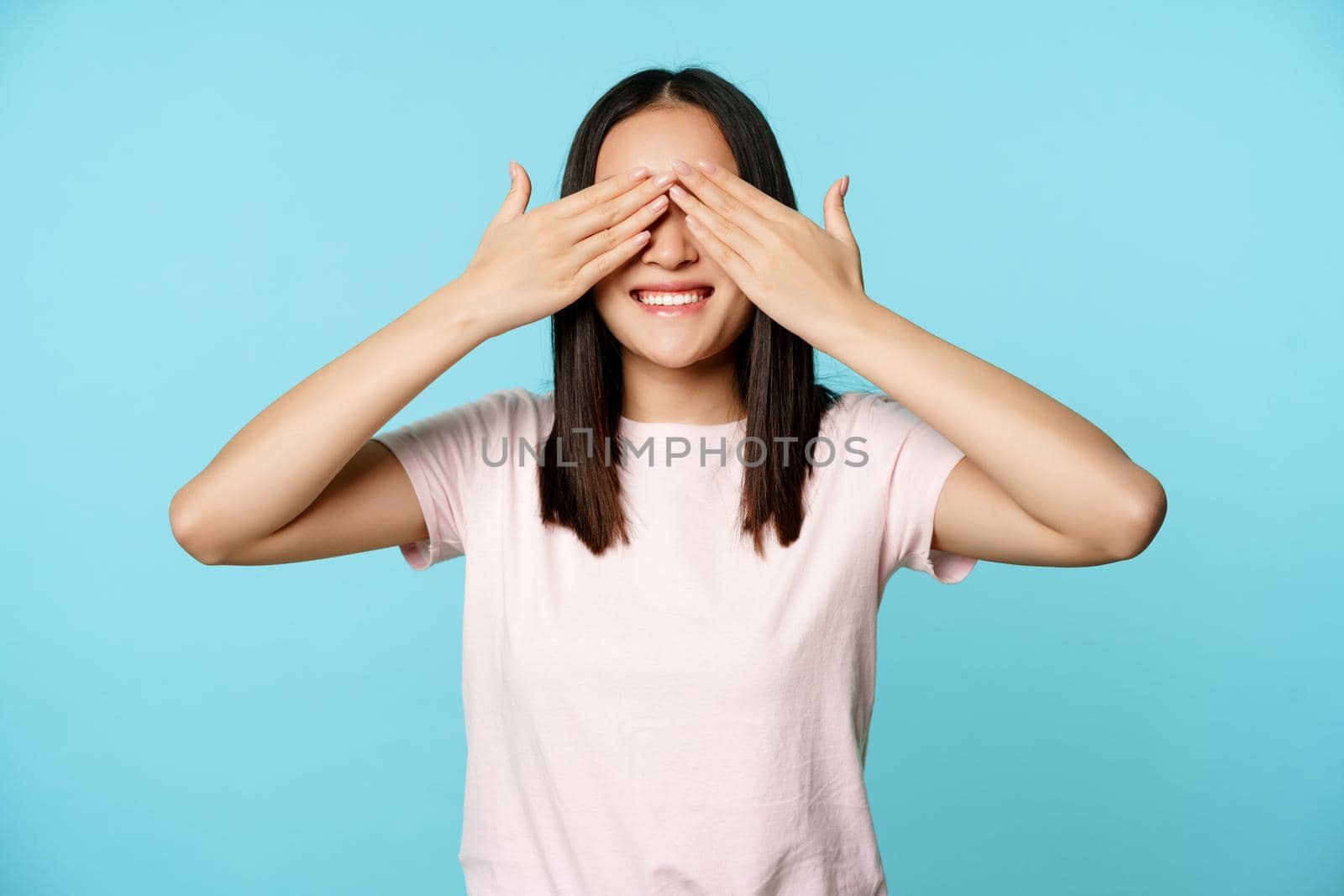 Happy smiling asian girl covers ears and waits for surprise, standing in t-shirt over blue background. Copy space