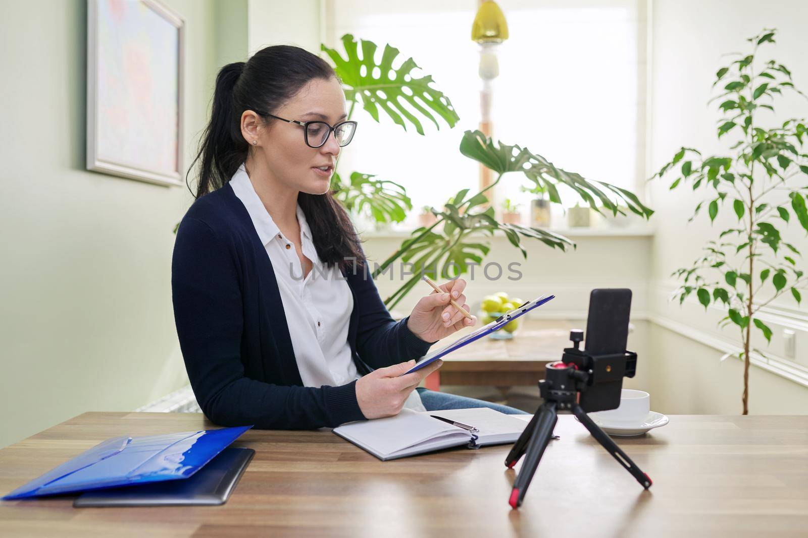 Business woman looking in smartphone webcam on tripod at home. Female talking online recording video blog, consulting finance, mentoring teaching training remotely, helping psychologist, distance work