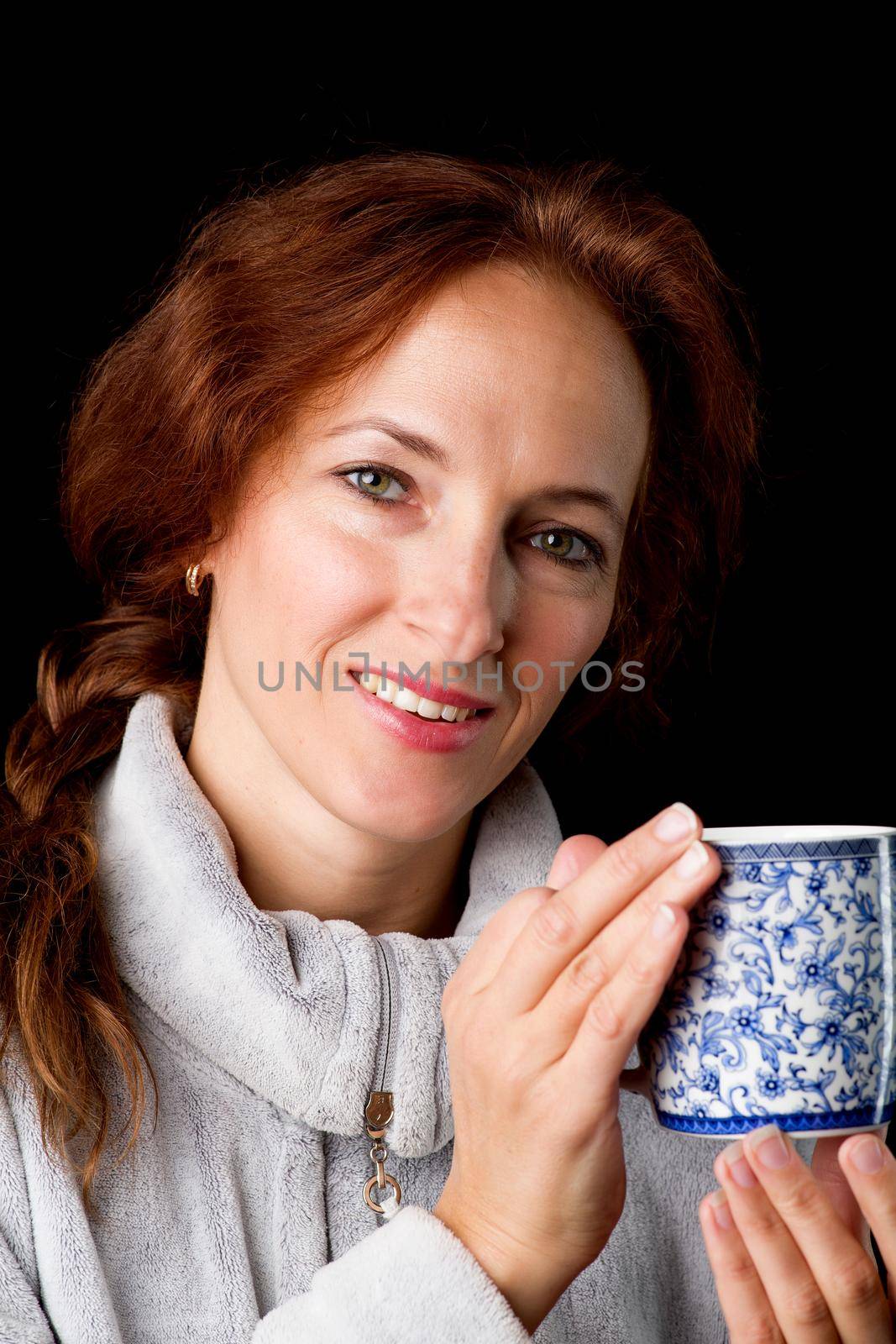 Woman holding mug in her hands. Cheerful brown haired woman in warm sweatshirt posing with cup of coffee or tea against black background. Female person enjoying of hot drink in the morning